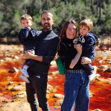 L'image représente une famille de quatre personnes debout devant un plan d'eau brun rougeâtre, avec des arbres visibles à l'arrière-plan. La famille est vêtue de vêtements sombres.