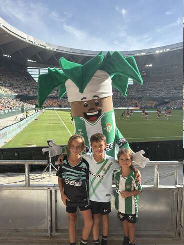 Tres niños con camisetas del Real Betis posan felices con la alegre mascota del equipo, una palmera, en un estadio de fútbol soleado y abarrotado durante un partido.