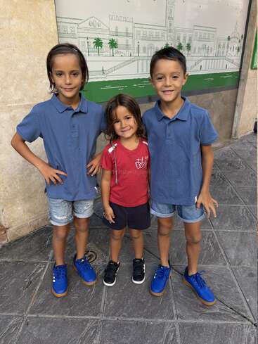 Tres niños posan y sonríen al aire libre, de pie sobre el pavimento de baldosas. Dos niños mayores llevan camisa azul y zapatillas de deporte, mientras que el más pequeño lleva camisa roja y zapatos negros.