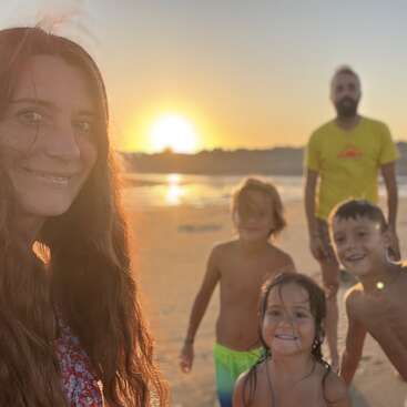 Una mujer, un hombre y tres niños sonríen en una playa durante la puesta de sol. La cálida luz del sol crea un resplandor dorado, capturando un alegre momento familiar junto al mar.