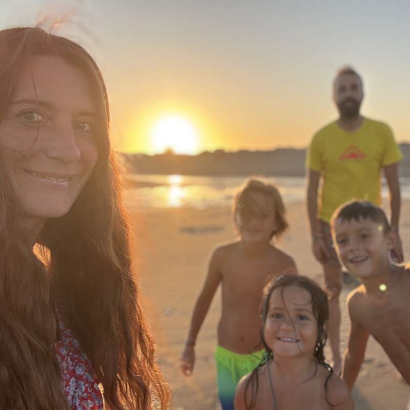 Una mujer, un hombre y tres niños sonríen en una playa durante la puesta de sol. La cálida luz del sol crea un resplandor dorado, capturando un alegre momento familiar junto al mar.