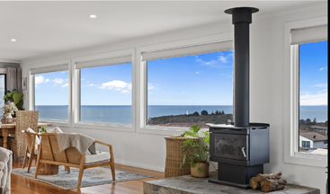 Dieses helle Wohnzimmer besticht durch große Fenster mit Meerblick, einen modernen Holzofen, gemütliche Sitzgelegenheiten, geflochtene Möbel, Zimmerpflanzen und Holzböden, die für eine ruhige Atmosphäre sorgen.