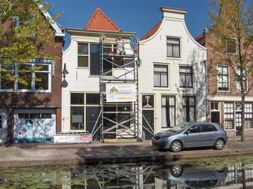The image depicts a white building with a red roof, featuring a man on scaffolding, a silver car, and a canal in front, set against a clear blue sky.