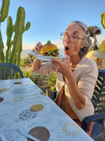 A woman with gray hair and large glasses excitedly holds a plate with a hamburger. She sits outdoors by a table, surrounded by cacti, enjoying sunny weather.