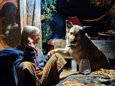 Una mujer está sentada fuera por la noche, con una porción de pizza en la mano y acariciando a un perro grande y peludo. Parecen felices. Sobre la mesa hay pizza y copas de vino.