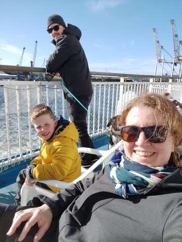 Une famille souriante profite d'une journée ensoleillée sur un ferry. La mère et le fils se détendent sur des chaises, tandis que le père se tient à proximité. Grues en arrière-plan.