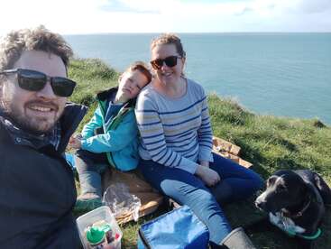 Une famille profite d'un pique-nique ensoleillé sur une falaise herbeuse au bord de la mer, souriant avec son chien, entouré de snacks et d'une eau d'un bleu éclatant.