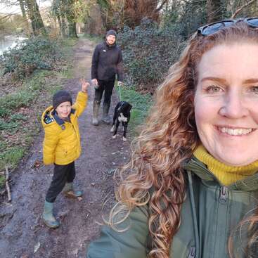 A woman with curly hair takes a selfie on a woodland path with a smiling child, a man, and a dog. Everyone appears happy and outdoorsy.
