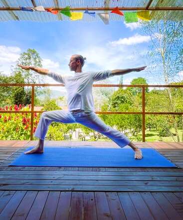 A person practices yoga on a blue mat outdoors, performing Warrior II pose. They wear white clothes with prayer flags above, surrounded by lush greenery and flowers.