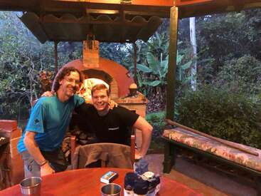 Two friends, smiling and embracing, pose happily in front of a lit brick oven outdoors. Surrounded by greenery, they enjoy a cozy, rustic patio evening.