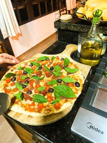 Une pizza fraîchement cuite sur une planche en bois est garnie de basilic, de tomates cerises, d'olives et de pepperoni. De l'huile d'olive et des ustensiles de cuisine se trouvent à proximité.