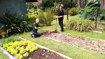 Deux personnes jardinent ensemble à côté d'une maison. L'une est agenouillée et souriante, l'autre est debout et tient une pelle. Il y a des plantes vertes et un potager.