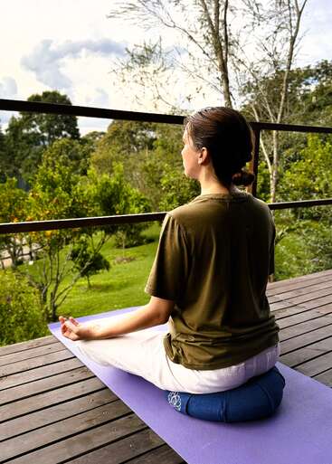 Une femme est assise en tailleur sur un tapis de yoga violet et médite paisiblement sur une terrasse en bois donnant sur un jardin verdoyant, luxuriant et tranquille, parsemé d'arbres.