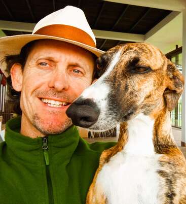 Un homme portant un chapeau blanc et une veste verte sourit en prenant un selfie avec un chien bringé à l'intérieur, capturant un moment chaleureux et heureux ensemble.