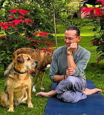 Un homme est assis en tailleur sur une natte à l'extérieur, souriant à un chien à côté de lui. Un autre chien se tient derrière, entouré d'une végétation luxuriante et de fleurs rouges.