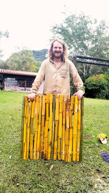 Un homme souriant aux cheveux bouclés se tient dehors, sur l'herbe, tenant un panneau fait de tiges de bambou jaunes. Derrière lui, on aperçoit des arbres, une maison et des montagnes.
