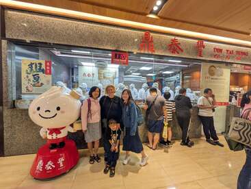 Um grupo de pessoas posa do lado de fora do restaurante Din Tai Fung, ao lado de um mascote de bolinho de massa. Atrás deles, chefs com uniformes brancos preparam alimentos em uma cozinha visível.