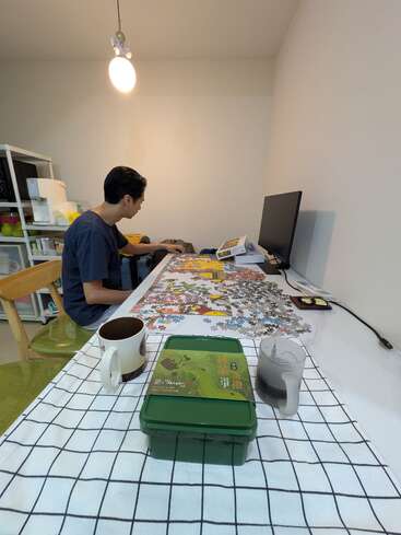 Uma pessoa está sentada em uma mesa trabalhando em um grande quebra-cabeça, com bebidas, uma caixa verde e um computador em uma sala aconchegante e bem iluminada.
