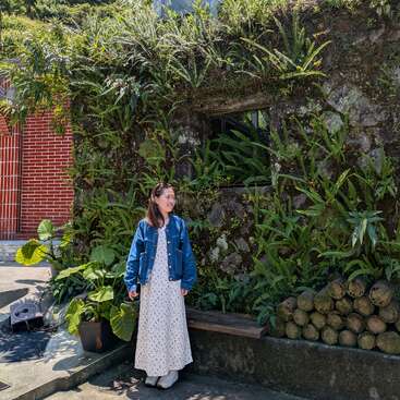 Uma mulher de vestido branco e jaqueta azul está ao lado de uma exuberante parede de pedra coberta de musgo com samambaias, perto de troncos empilhados e vasos de plantas, ao ar livre.