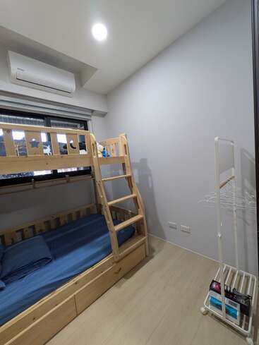 Esta imagem mostra um quarto minimalista com beliches de madeira, roupa de cama azul, uma janela, ar-condicionado, piso de madeira clara e um simples cabideiro branco.