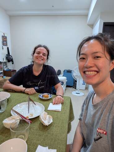Deux amis souriants sont assis à une table avec des assiettes terminées et un dessert. La salle confortable présente un décor simple, une nappe verte et une atmosphère détendue et amicale.