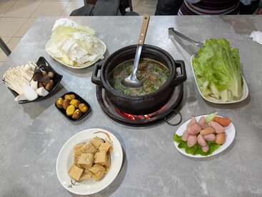 Una comida de olla caliente se prepara con una olla a fuego lento con caldo, una variedad de setas, verduras de hoja verde, tofu, carnes, castañas y verduras dispuestas sobre una mesa.