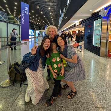 Vier Menschen posieren fröhlich zusammen in einem Flughafenterminal in der Nähe des Abflugbereichs, umgeben von Geschäften und hellen Lichtern. Eine Person zeigt ein Friedenszeichen.