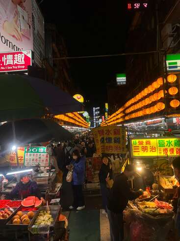 Um mercado noturno cheio de gente, com lanternas amarelas brilhantes, barracas lotadas vendendo comida e frutas, pessoas fazendo compras e placas em chinês, criando uma atmosfera animada.