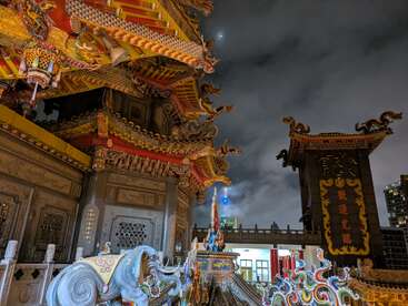 Un templo chino bellamente ornamentado por la noche, ricamente decorado con dragones coloridos y tallas intrincadas, iluminado bajo un cielo nublado con una vista lejana de la ciudad.