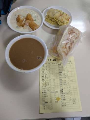 Esta imagen muestra un desayuno taiwanés: leche de soja, youtiao en leche de soja salada, un rollito de tortilla de huevo, un sándwich y una hoja amarilla con el pedido sobre la mesa.