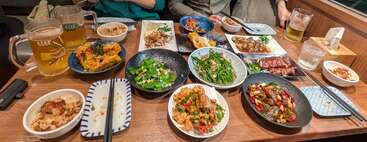 Una mesa repleta de diversos platos asiáticos, incluyendo verduras, carnes y arroz, acompañados de jarras de cerveza y bebidas, preparada para que varias personas disfruten de la cena.