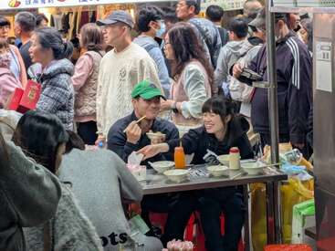 Una animada escena de mercado nocturno con una multitud de gente. Dos personas se sientan en un puesto de comida, compartiendo comida y risas, rodeadas de energía y animadas conversaciones.