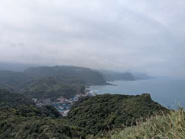 Esta imagen muestra exuberantes colinas verdes con vistas a una pequeña ciudad costera junto al mar, con montañas que se desvanecen en la brumosa y nublada distancia bajo un cielo gris.