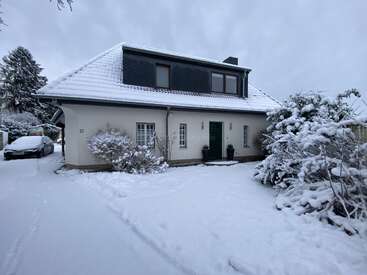 Ein zweistöckiges Haus mit schneebedecktem Dach und Garten, umgeben von schneebedeckten Büschen, steht unter einem bewölkten Winterhimmel. Draußen steht ein Auto.