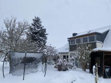 Ein modernes Haus steht in einem verschneiten Garten, mit Bäumen und Büschen, die ganz mit Schnee bedeckt sind. Im Hof steht ein Trampolin, das auch total zugeschneit ist.