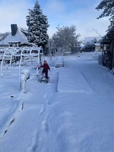 Ein Kind in einem roten Schneeanzug läuft durch einen verschneiten Hof mit Häusern und Bäumen, die ganz in Weiß sind, unter einem strahlend blauen Winterhimmel.