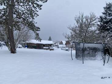 Eine verschneite Gartenszene mit Trampolin, kahlen Bäumen und Schuppen. Der Schnee bedeckt den Boden und die Dächer und sorgt überall für eine ruhige, winterliche und friedliche Atmosphäre.