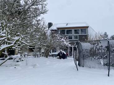Ein modernes Haus steht umgeben von tiefem Schnee, mit schneebedeckten Bäumen, Gartenmöbeln und einem Trampolin im Hinterhof und schafft so eine friedliche Winterlandschaft.