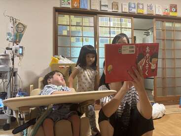 Uma mulher lê um livro vermelho para duas crianças em uma sala aconchegante. Você pode ver equipamentos médicos ao lado do menino em uma cadeira, sugerindo um ambiente de cuidados.