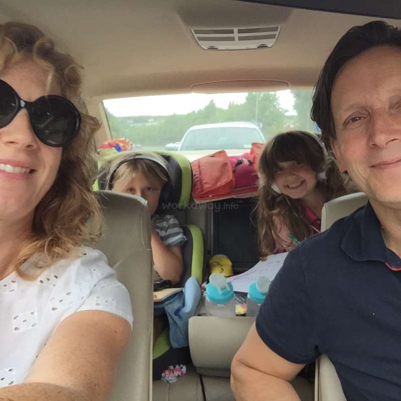 Das Bild zeigt eine vierköpfige Familie in einem Auto, mit zwei Kindern auf dem Rücksitz, die in die Kamera lächeln, auf einer Autoreise.