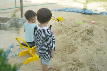 Duas crianças pequenas brincam juntas em um playground de areia. Uma delas senta-se em um brinquedo amarelo de escavação, enquanto a outra fica por perto, ambas concentradas na pilha de areia.