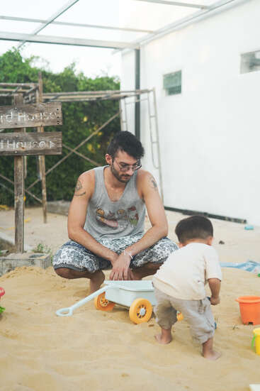 Um homem e uma criança estão brincando juntos em uma área externa de areia, cercados por brinquedos. A atmosfera parece relaxada e divertida, com vegetação ao fundo.