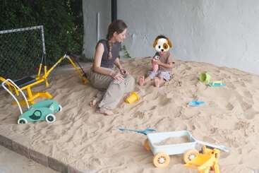 Uma mulher e uma criança com um filtro de rosto de cachorro estão sentadas em uma caixa de areia, cercadas por vários brinquedos coloridos, participando de atividades lúdicas e criativas.