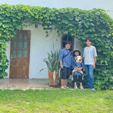 Três pessoas posam felizes em frente a uma casa charmosa coberta de vinhas verdes exuberantes com flores roxas. O rosto de uma pessoa está coberto por um emoji de cachorro.