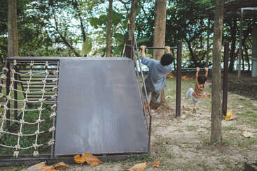 Deux enfants jouent en plein air sur un terrain de jeu, se balançant sur des barres horizontales entourées d'arbres, de terre et de feuilles mortes, avec des équipements d'escalade visibles à proximité. C'est une journée ensoleillée.