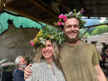 Duas pessoas sorridentes usam coroas de flores coloridas, bem juntinhas debaixo de um abrigo rústico. A vegetação exuberante, o clima relaxado e outras pessoas ao fundo deixam o ambiente ainda mais festivo.