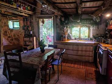Essa cozinha rústica e aconchegante tem vigas de madeira, paredes de pedra, móveis ecléticos, uma mesa de jantar com flores, prateleiras abertas e luz do sol entrando pelas janelas e pela porta aberta.