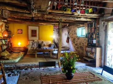 Esta acogedora sala de estar rústica cuenta con vigas de madera, paredes de piedra, una decoración colorida, estanterías, una guitarra, cojines, iluminación cálida y una mesa con un arreglo floral fresco.