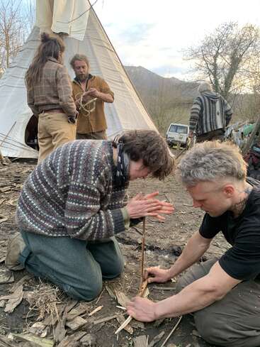Dos personas están arrodilladas en el suelo, haciendo fuego con un taladro de arco. Detrás de ellas, otras tres conversan cerca de un tipi, con montañas y árboles al fondo.