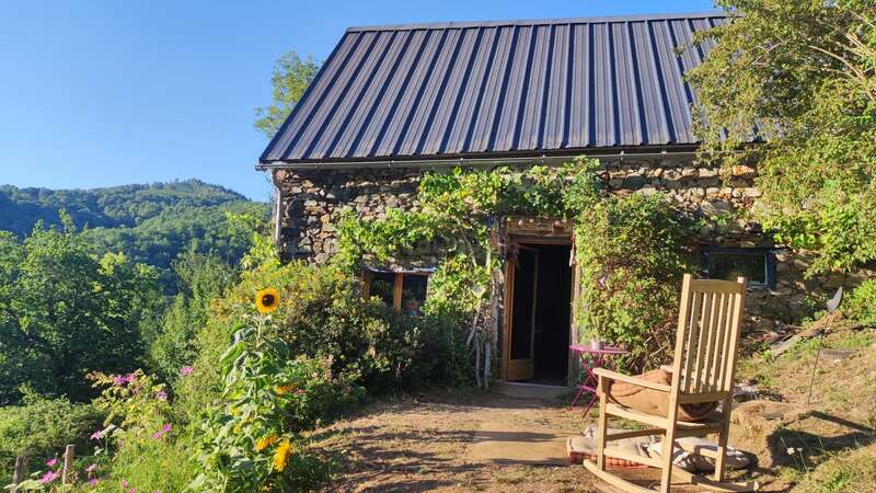 Uma charmosa cabana de pedra com telhado de metal fica no meio de uma vegetação exuberante e flores silvestres. Uma cadeira de balanço de madeira fica de frente para colinas vibrantes sob um céu azul claro.